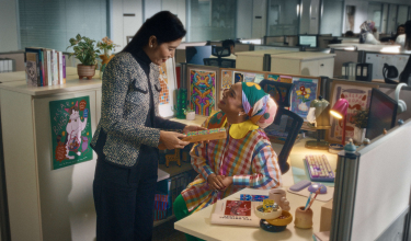 Office worker seated at desk decorated with local artworks engaged in conversation with her boss