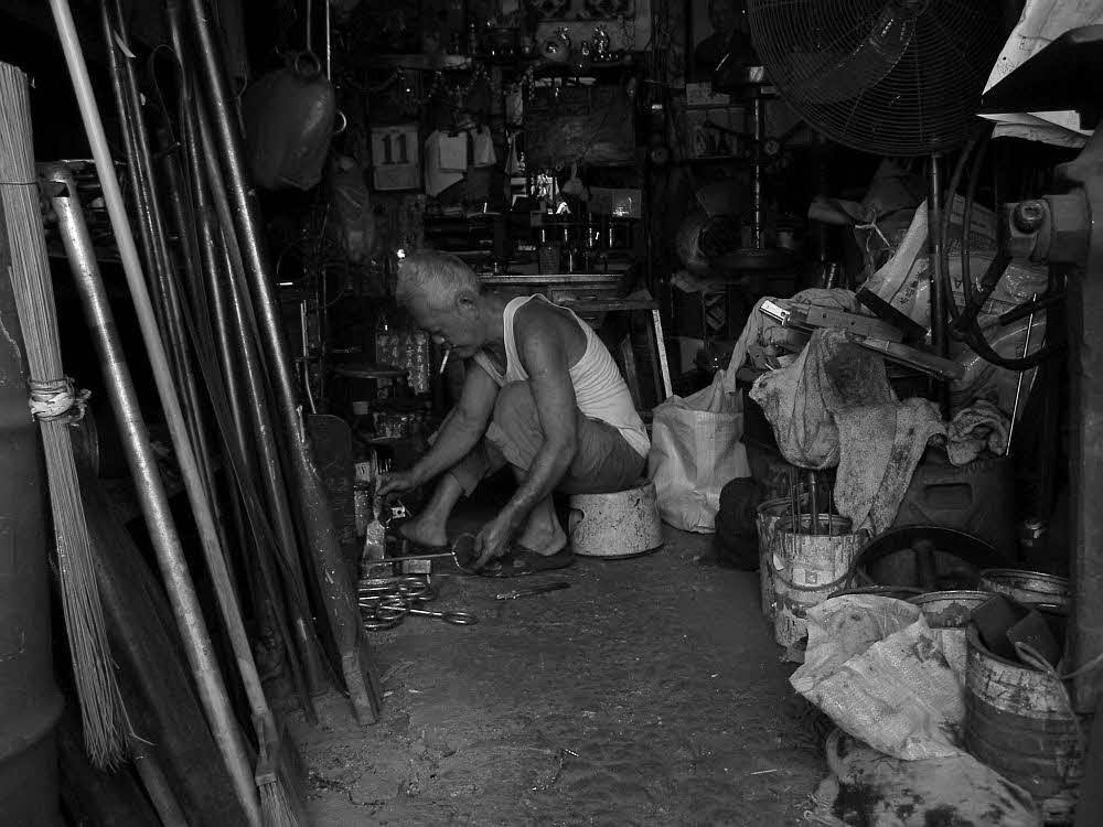 An old image of Sultan Gate Photo credit: Akai Chew A black-and-white photo of old Sultan Gate: an elderly man sits on a small stool, working with his hands on things on the ground. A Particular Place: Mapping Memories in Kampong Glam