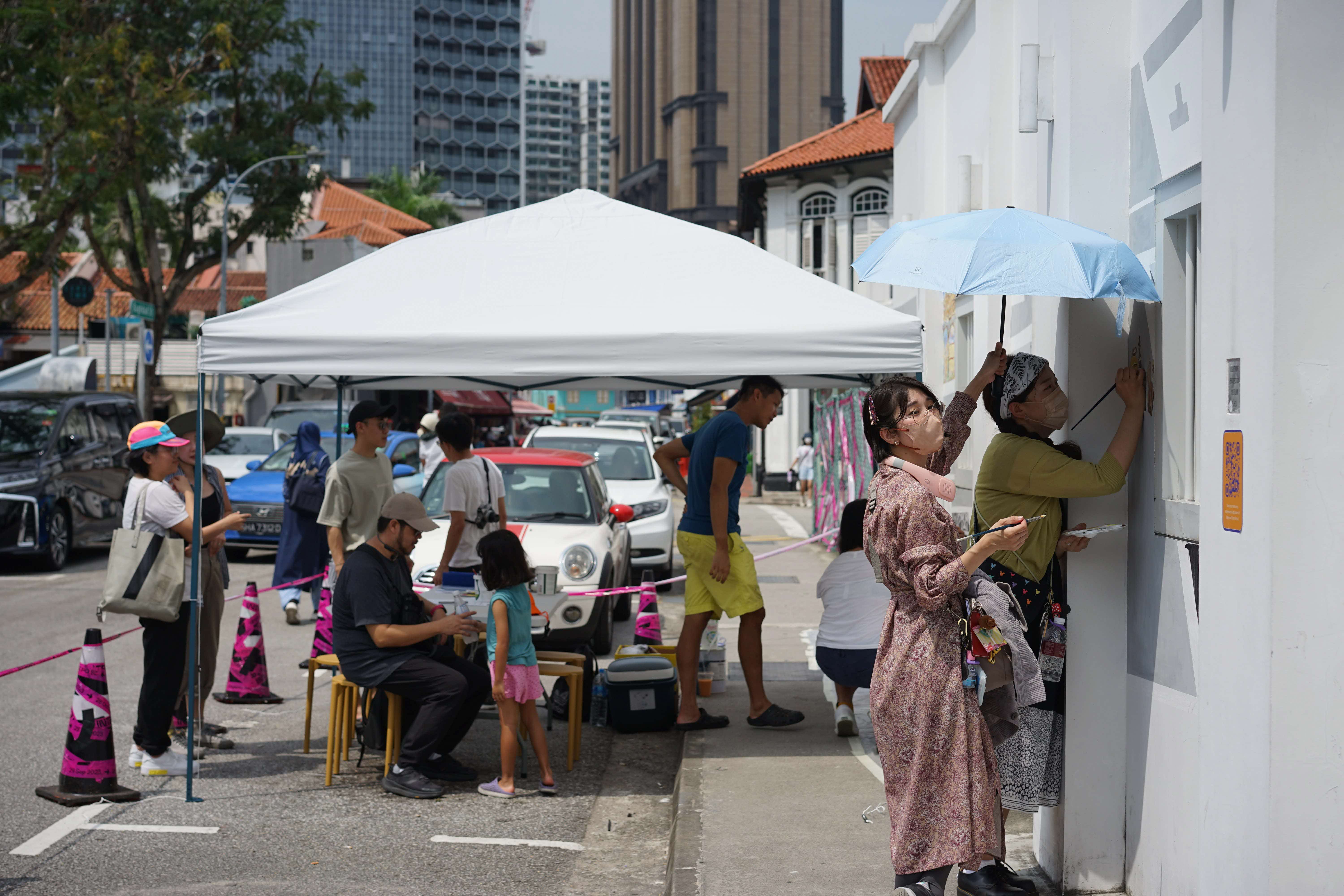 Volunteers and passersby adding their piece during co-creation sessions Photo credit: Singapore Archifest 2023 Curatorial Team. Members of the public gather at the sunny street of Sultan Gate. A few people are drawing on the wall of a building. A Particular Place: Mapping Memories in Kampong Glam