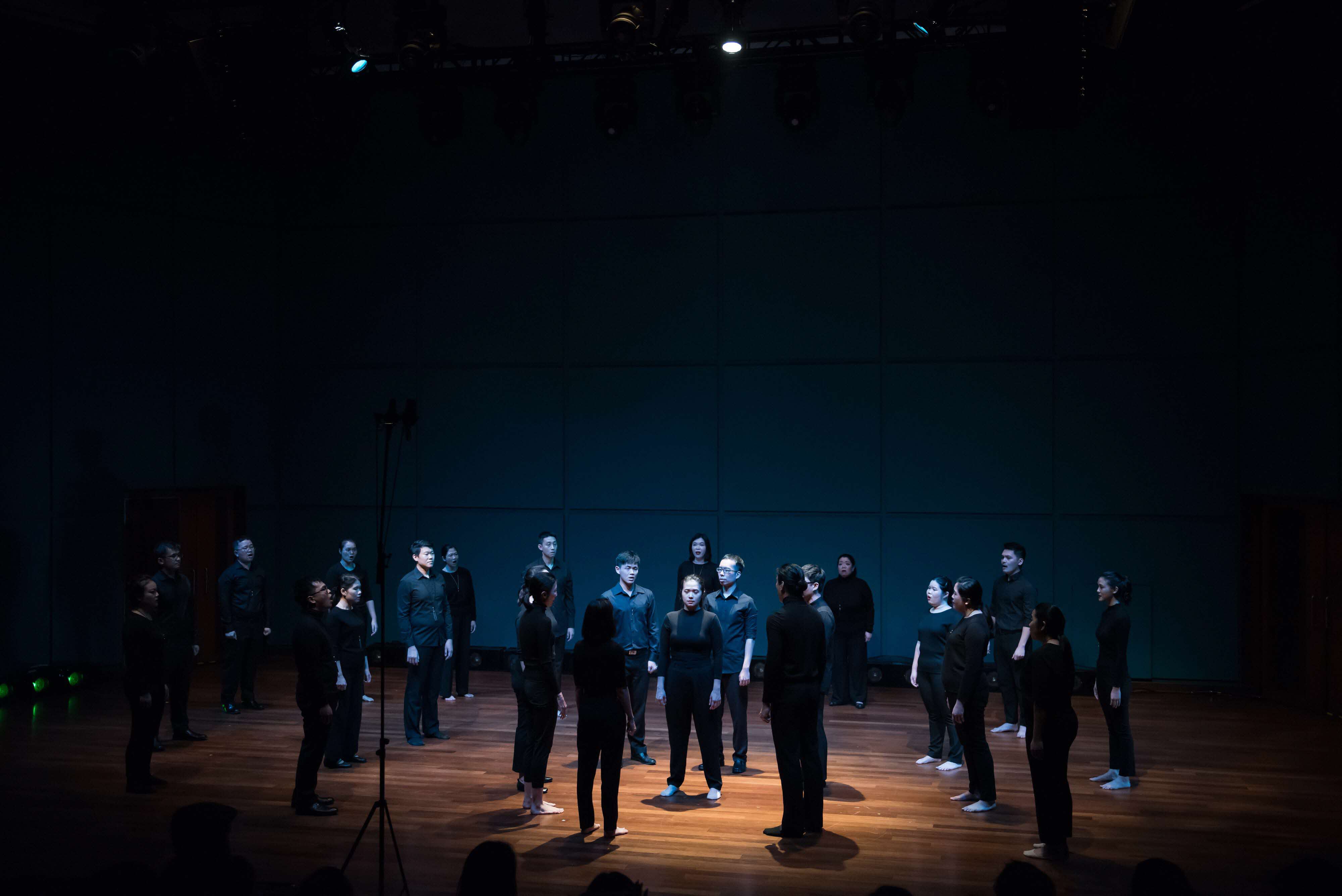 Multiple singers surround one central singer, bathed in a pale blue light.  Beyond Music: Singapore Youth Choir Ensemble Singers Explore the Visual and Aural World