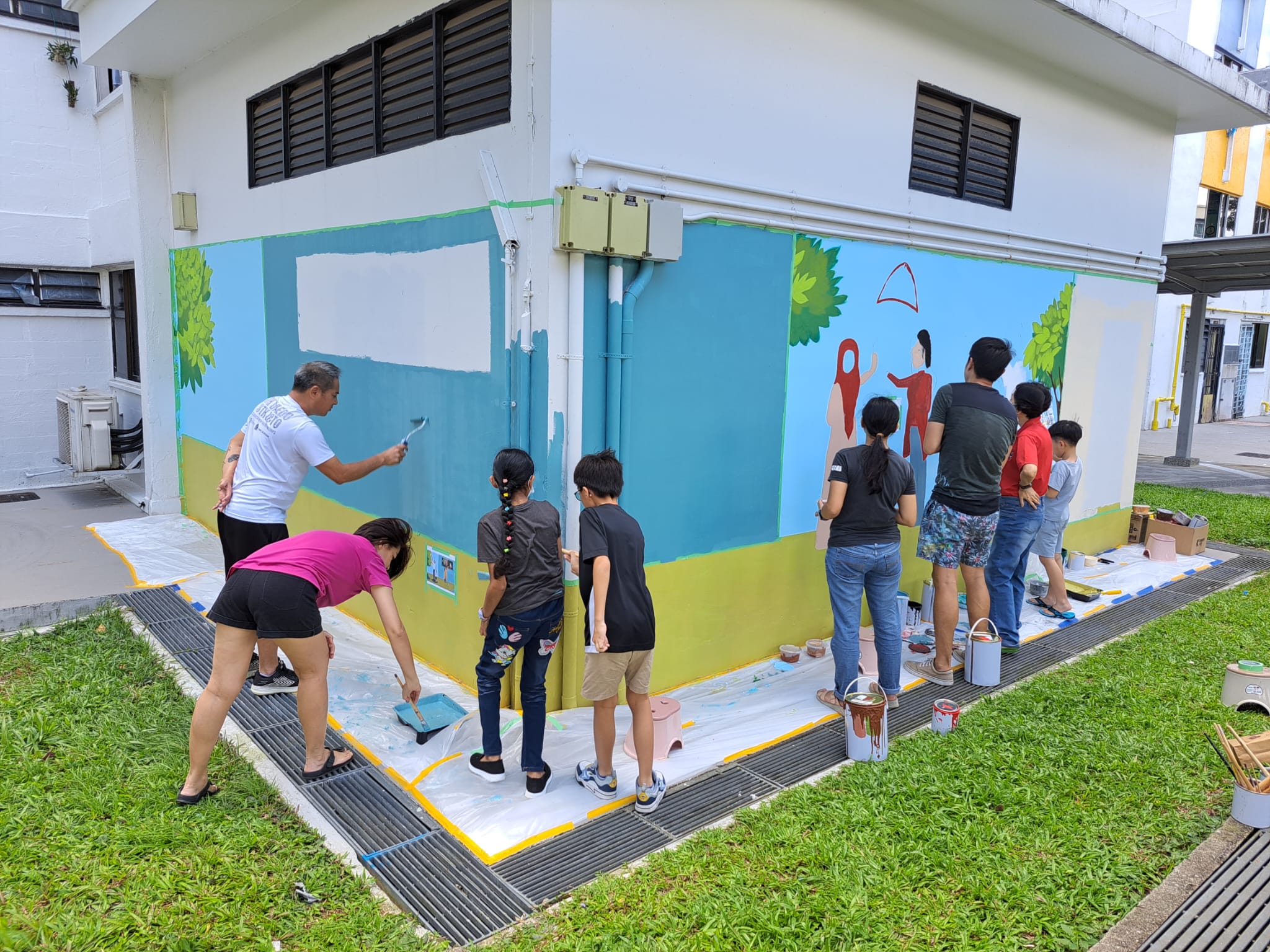 Members of the community work together on an outdoor wall mural, showing a stretch of blue sky