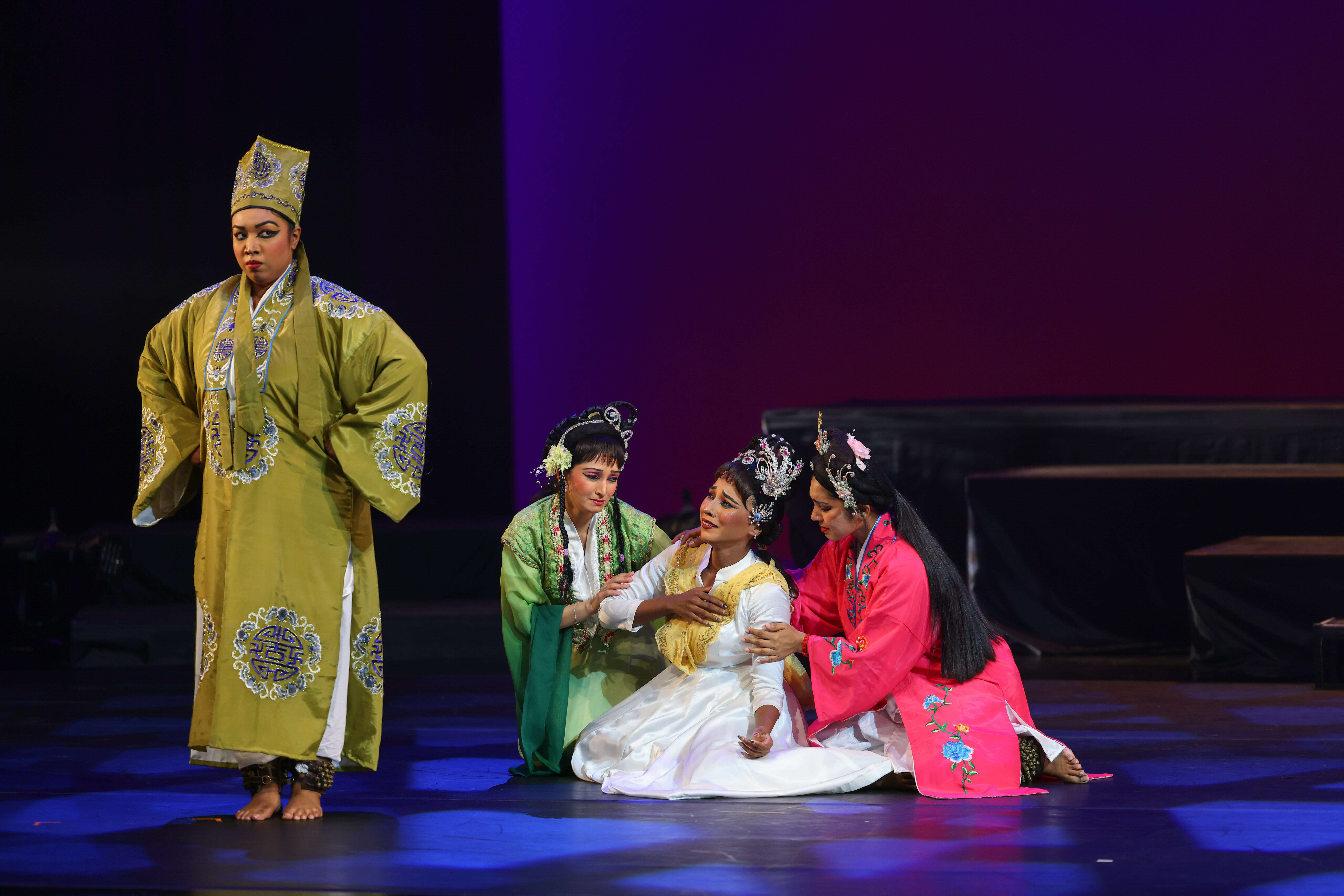 Performers on stage in traditional Chinese theatre costumes – yellow, green, and pink robes with ornate headdresses.  "Butterfly Lovers": A Timeless Tale Reimagined Through Generations