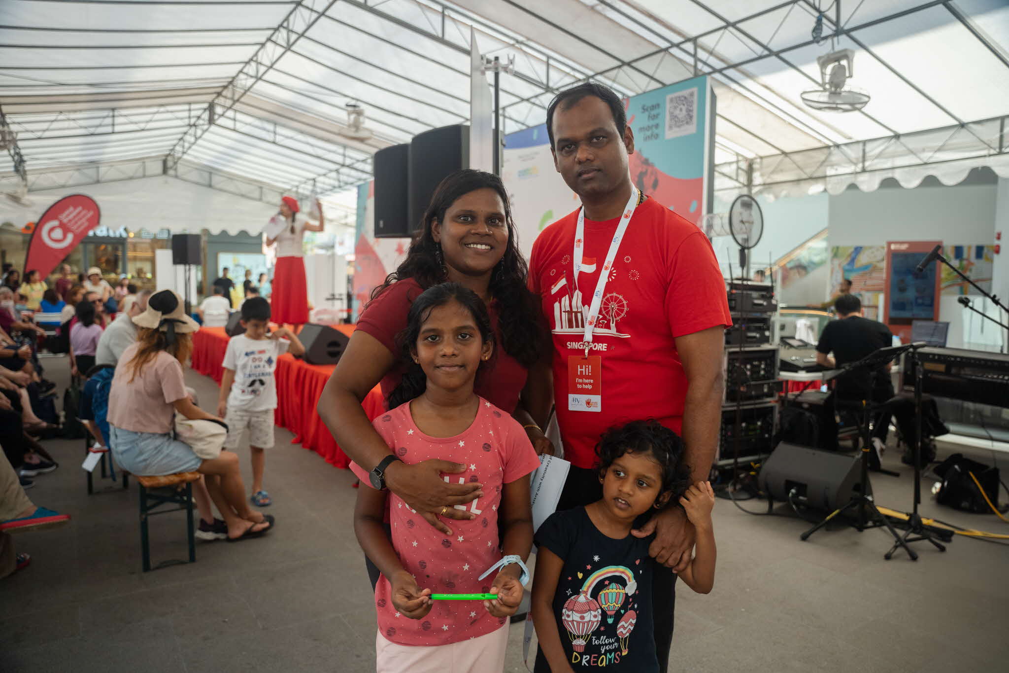 Krishnadevi and her husband enjoying an ArtsEverywhere@CDC programme at One Holland Village with their daughters. A family of four (two parents and two children) wearing red shirts posing for the camera in front of the ArtsEverywhere@CDC performance stage. Discover Joy Together: ArtsEverywhere@CDC — Creating Family Memories through Art