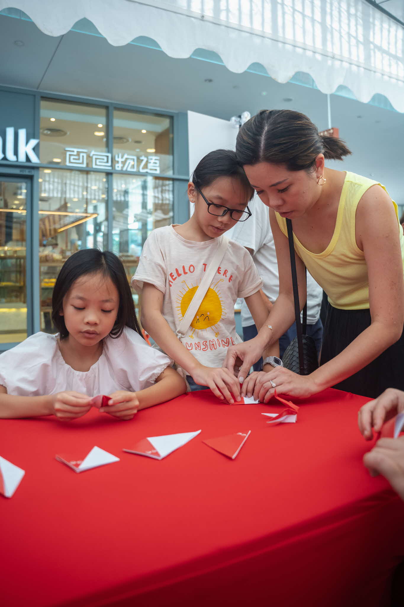 Yvonne, a mother, joining her daughters in the Crane Origami workshop at ArtsEverywhere@CDC. A mother and her two daughters engrossed in folding and making origami cranes at a red table. Discover Joy Together: ArtsEverywhere@CDC — Creating Family Memories through Art