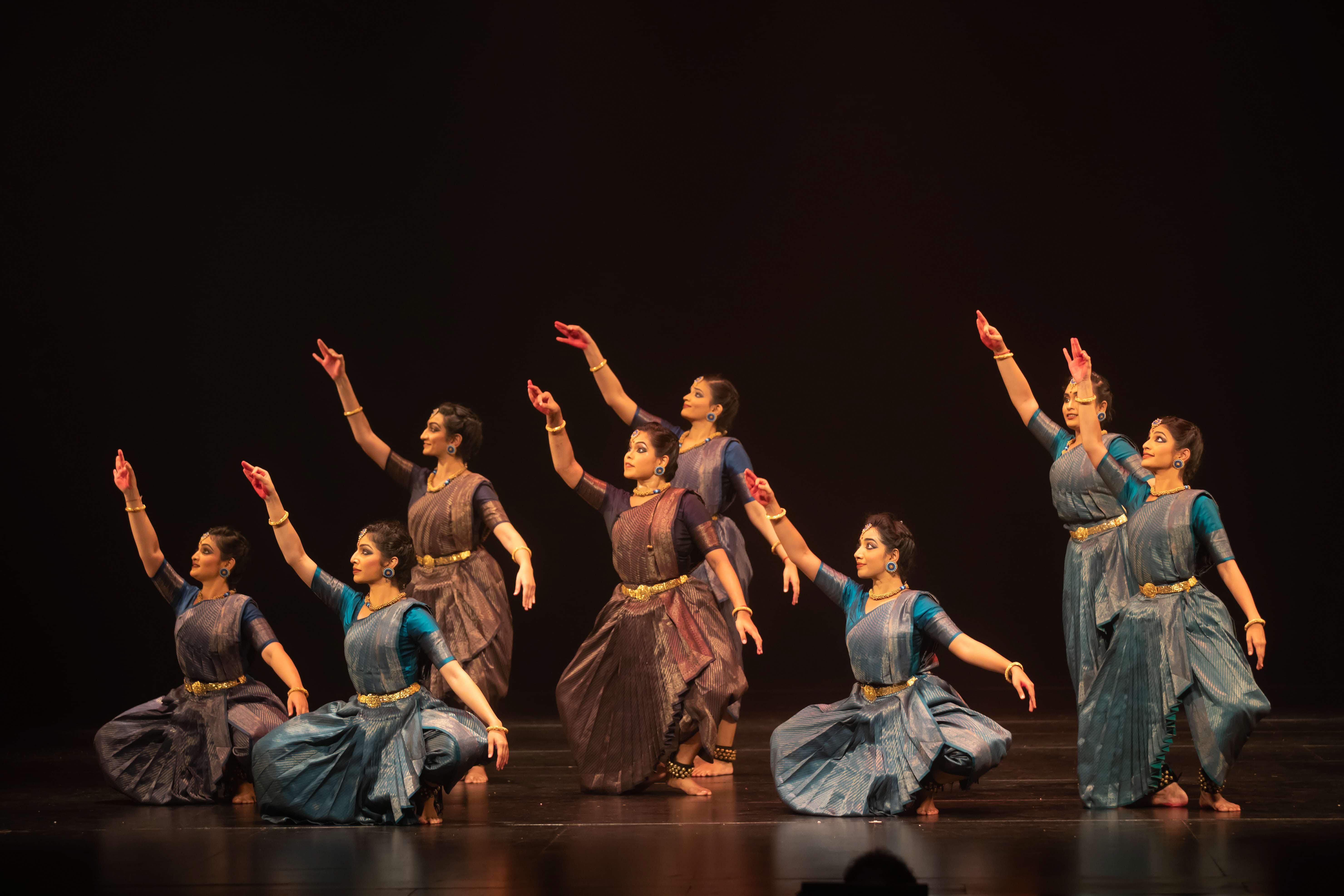 Dancers in mid-motion, showcasing their elegant lines for ‘ARISI:Rice’. Indian dancers performing on stage with arms raised in a synchronised pose.