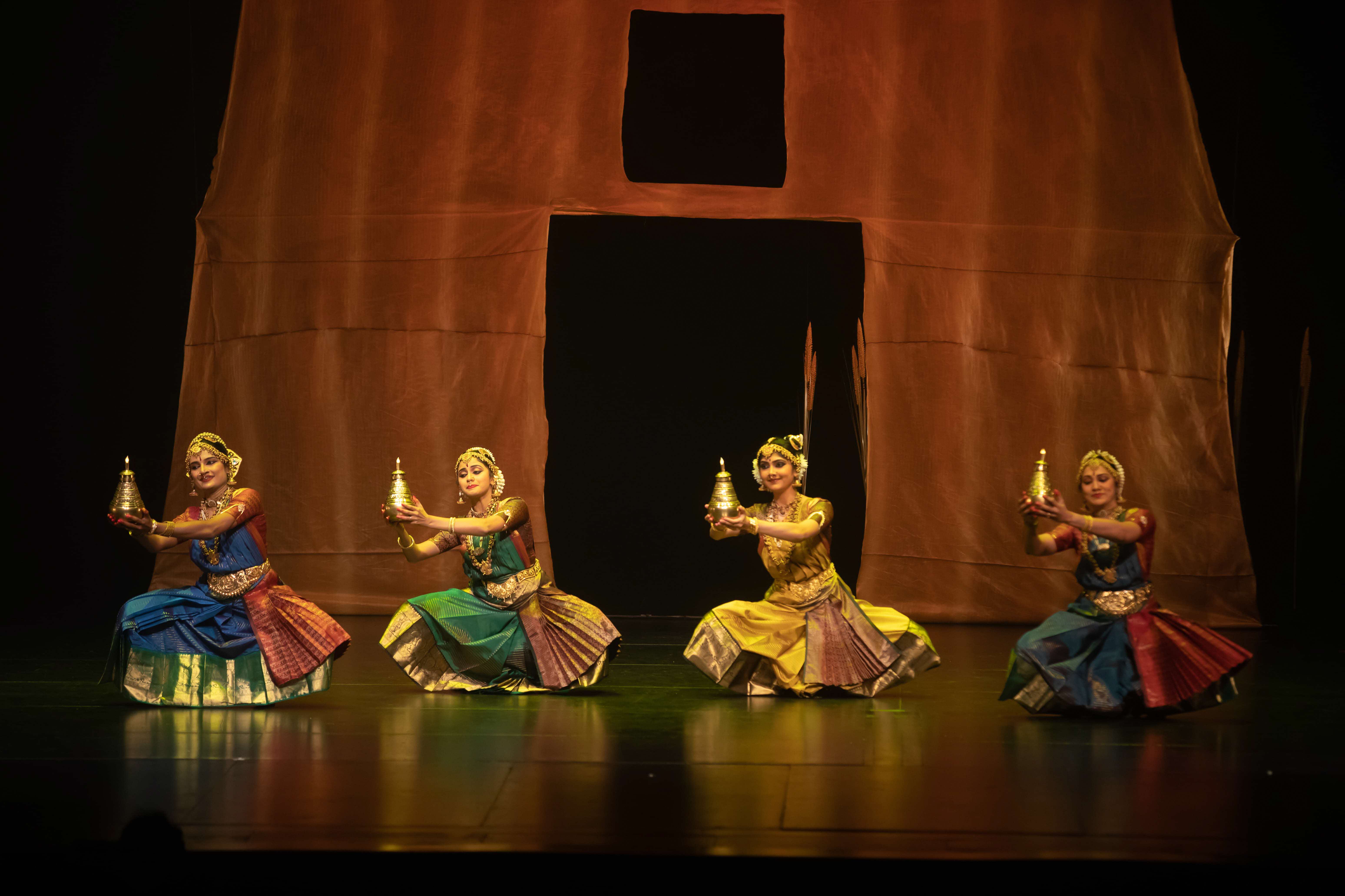 Dancers lined up in a row for ‘ARISI:Rice’. Four classical Indian dancers holding a golden ceremonial lamp, in coordinated dance poses.